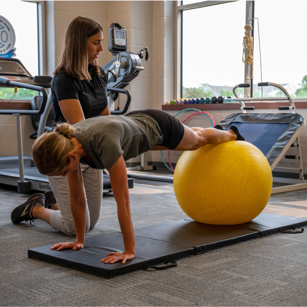 A woman in a plank position keeps her feet on an exercise ball. Her physical therapist assists her in the physical therapy clinic.