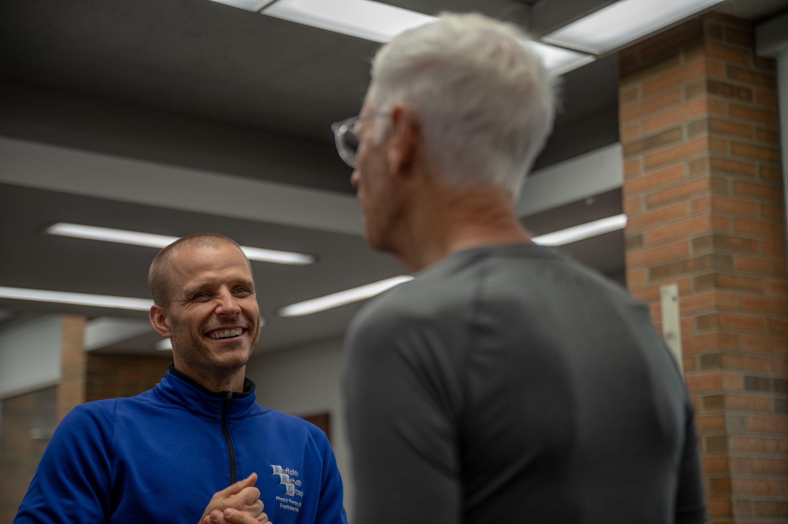 A man smiles as he speaks to another man. The smiling man is wearing a blue zip up sweater. The other man wears a gray shirt.