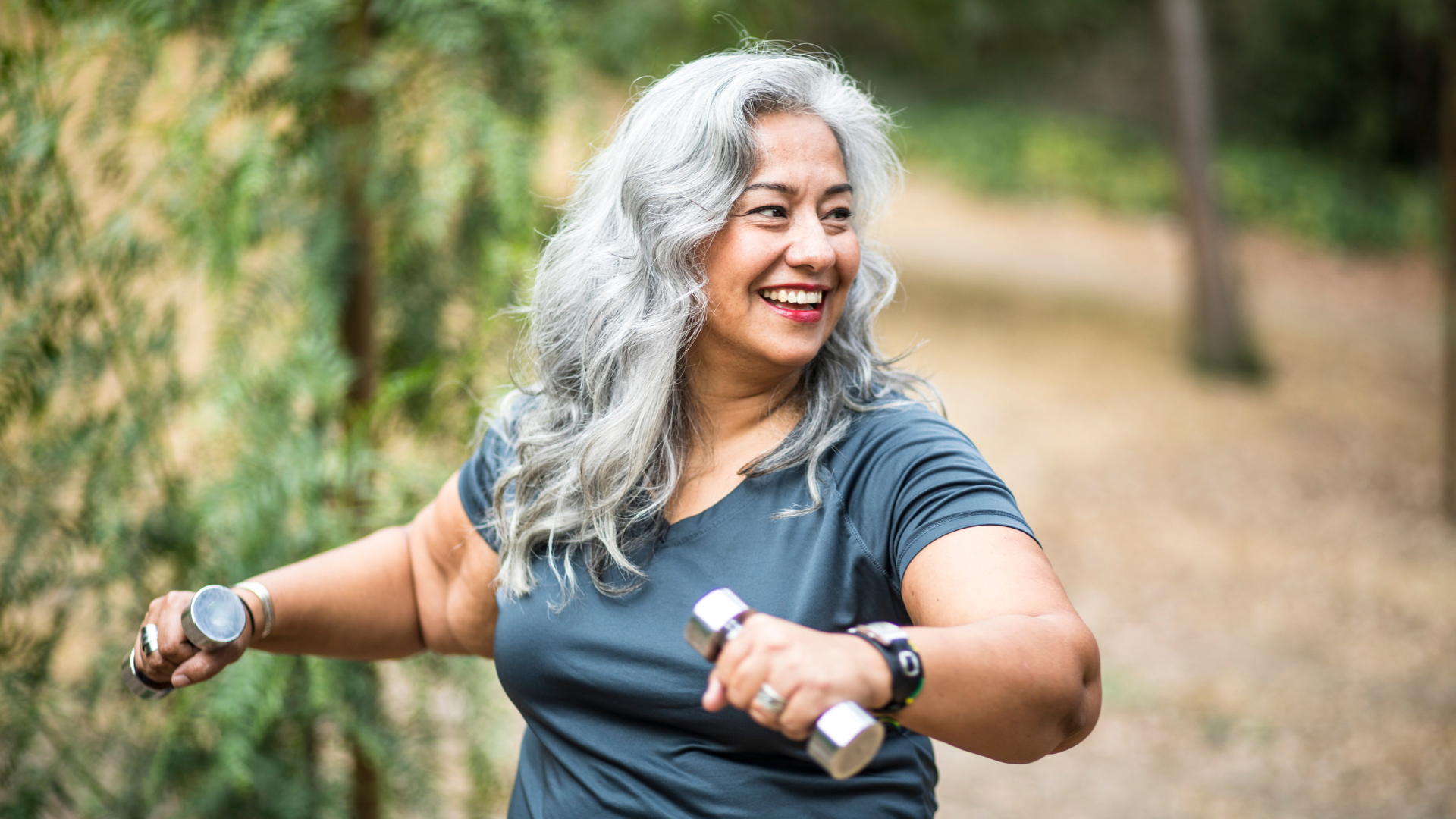 A woman with gray hair and a blue t-shirt smiles as she power walks with two small weights in her hands.